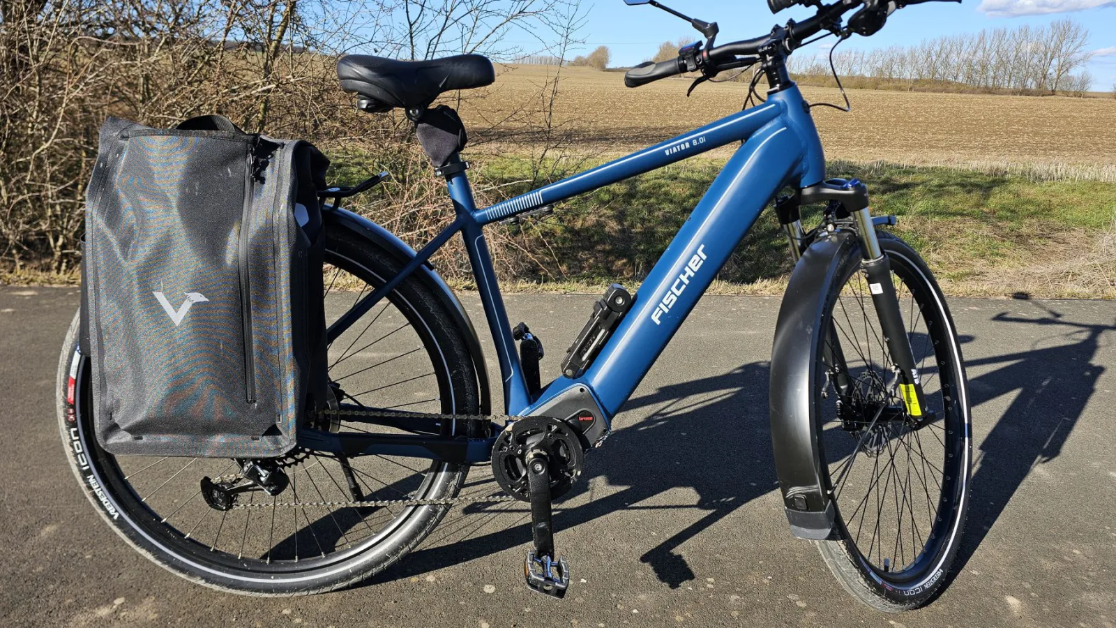 Blaues Fischer Viator Trekking-E-Bike mit Gepäckträgertasche am Feldrand.